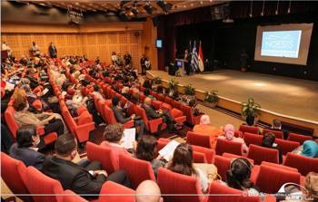 Many people sit on chairs in a theatre and watch the speech of the man on the podium
